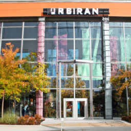 description: an image shows the exterior of urban outfitters' new store location at oxmoor center in louisville. the store has large glass windows showcasing trendy clothing displays. the entrance is adorned with colorful banners and a prominent logo. shoppers can be seen entering and exiting the store, carrying urban outfitters' signature shopping bags.