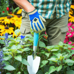 A person wearing gardening gloves and holding a small spade, standing in a garden filled with plants and flowers.