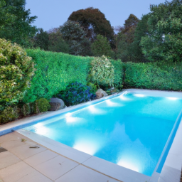 A backyard swimming pool with crystal blue water, surrounded by lush green trees and bushes. The pool has a white plaster finish and is illuminated by underwater lights, creating a peaceful and inviting atmosphere.