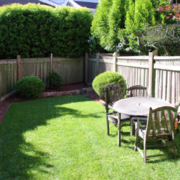 Description: A bright green lawn with a wooden fence in the background, a patio with a table and chairs, and a variety of plants and shrubs around the edges.